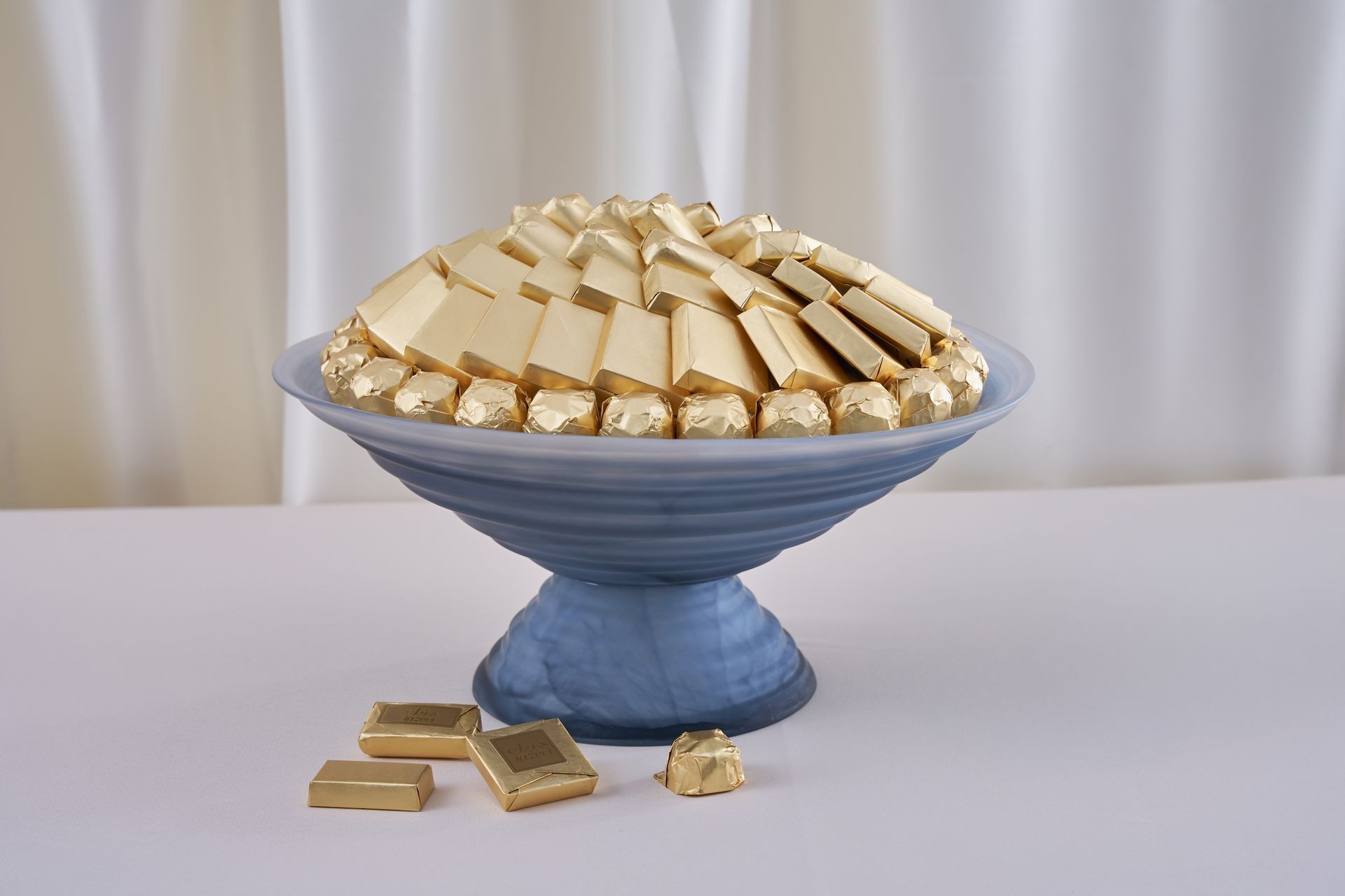 A blue glass bowl filled with assorted gold-wrapped chocolates placed on a white tablecloth.