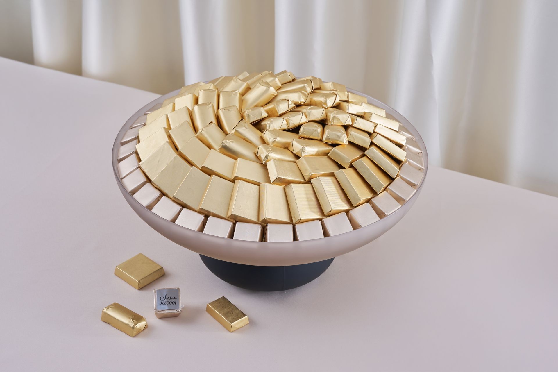 Circular display of gold and silver wrapped chocolates on a white stand with two pieces beside it.