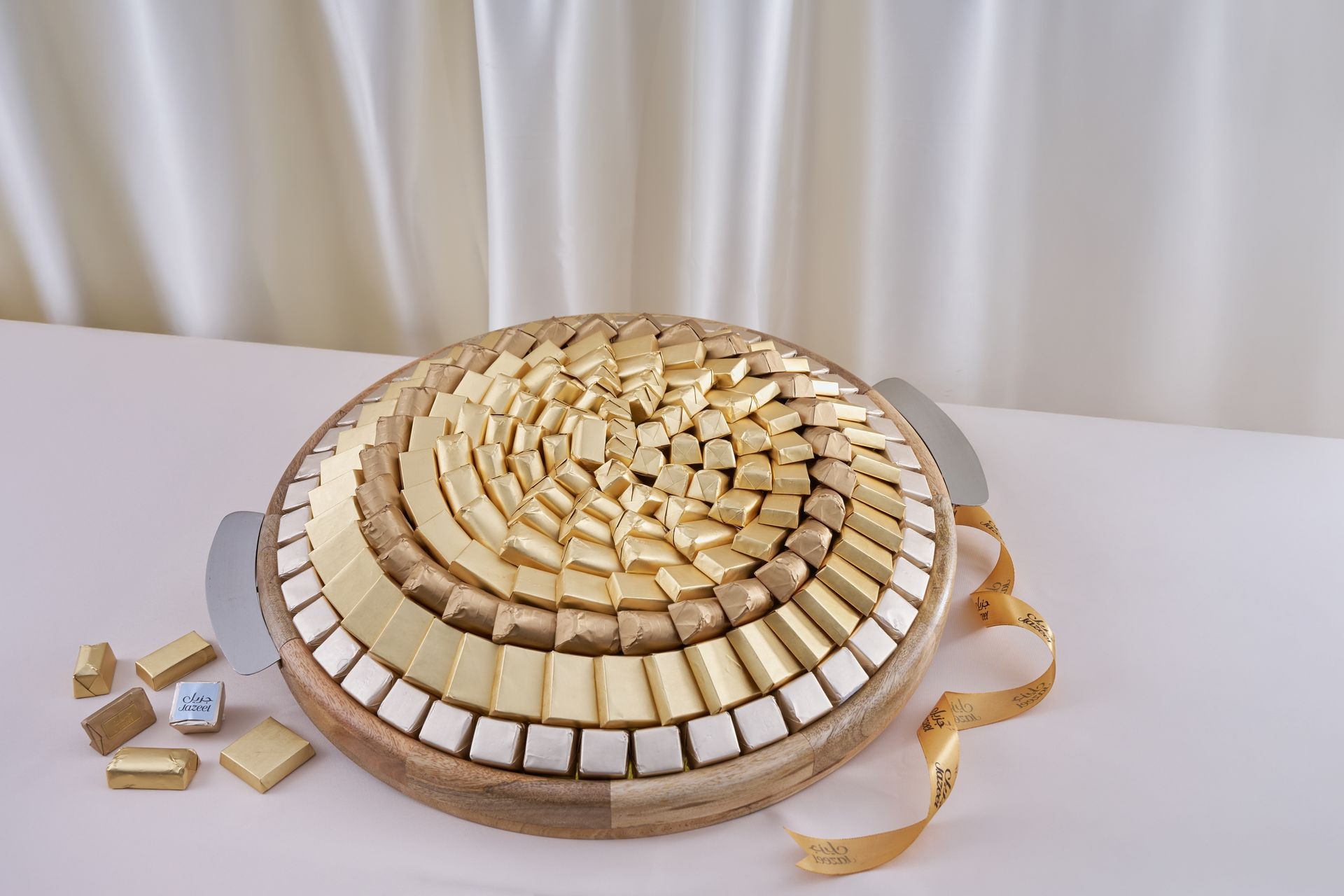 Round platter with neatly arranged layers of gold and silver wrapped chocolates, set on a white table.