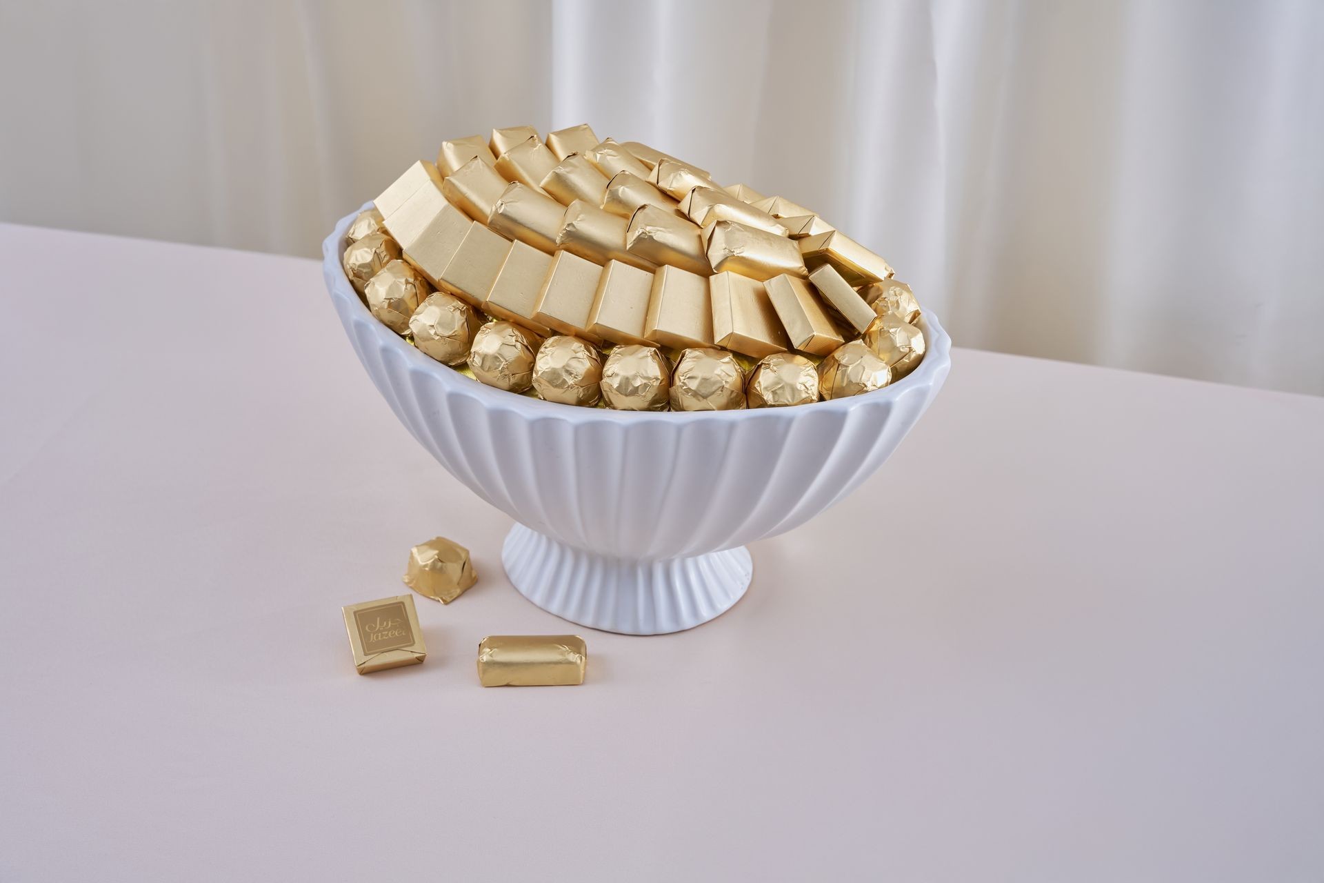White bowl filled with assorted gold-wrapped chocolates on a light pink surface.