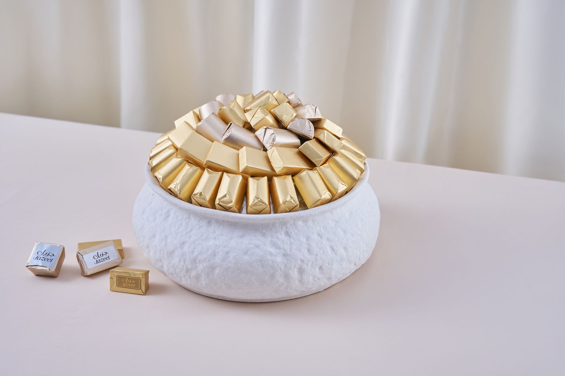 Elegant bowl filled with gold and silver wrapped chocolates, placed on a light beige table.