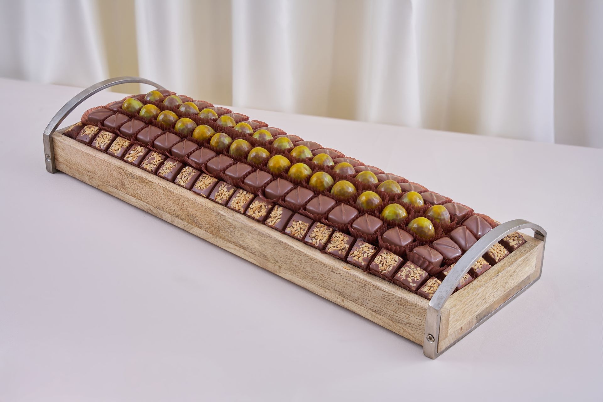 Wooden tray filled with rows of assorted gourmet chocolates on a light surface.