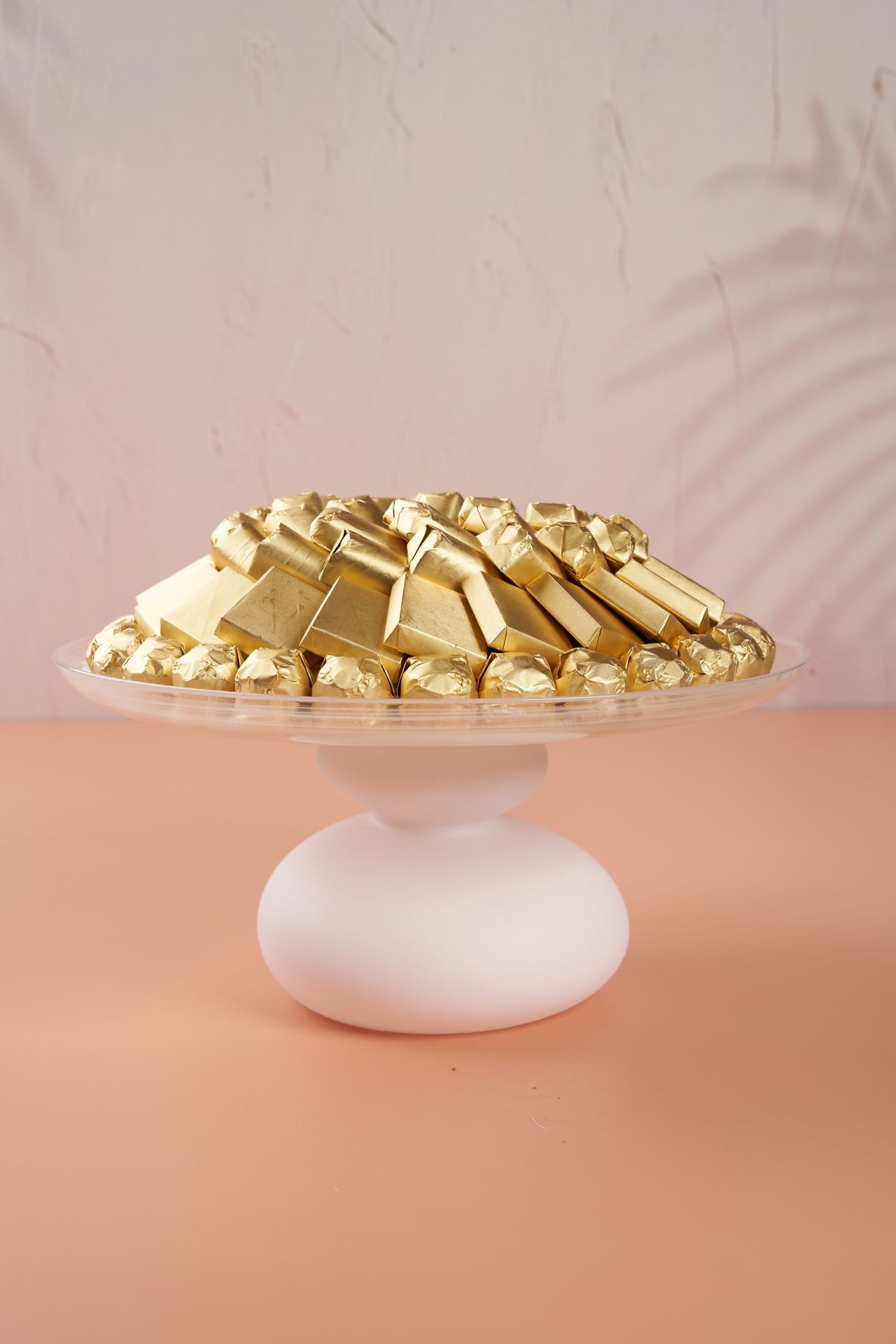 Gold-wrapped chocolates piled on a white cake stand against a pink background.