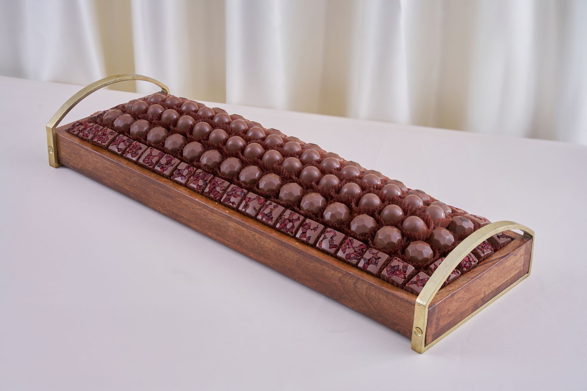 Wooden serving tray with rows of assorted chocolate bonbons on a white tablecloth.