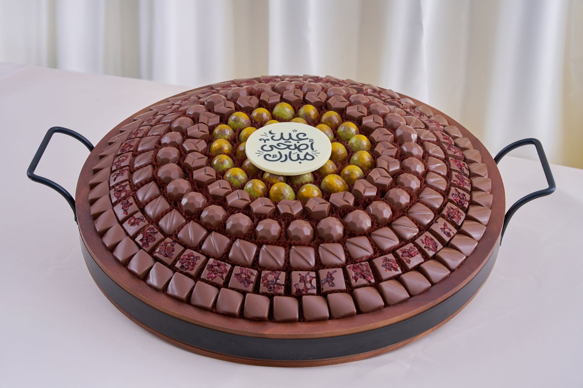 Large round tray with assorted chocolates and a decorative center piece on a white tablecloth.