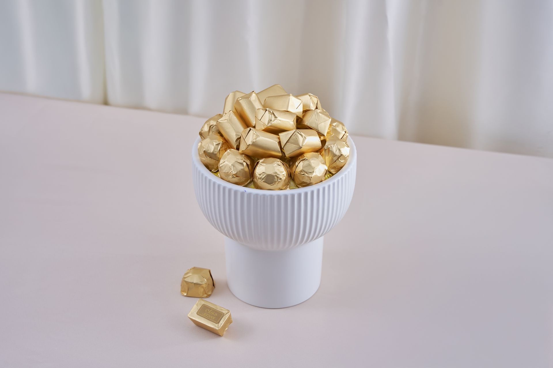White bowl filled with assorted chocolates wrapped in gold foil on a light-colored table.