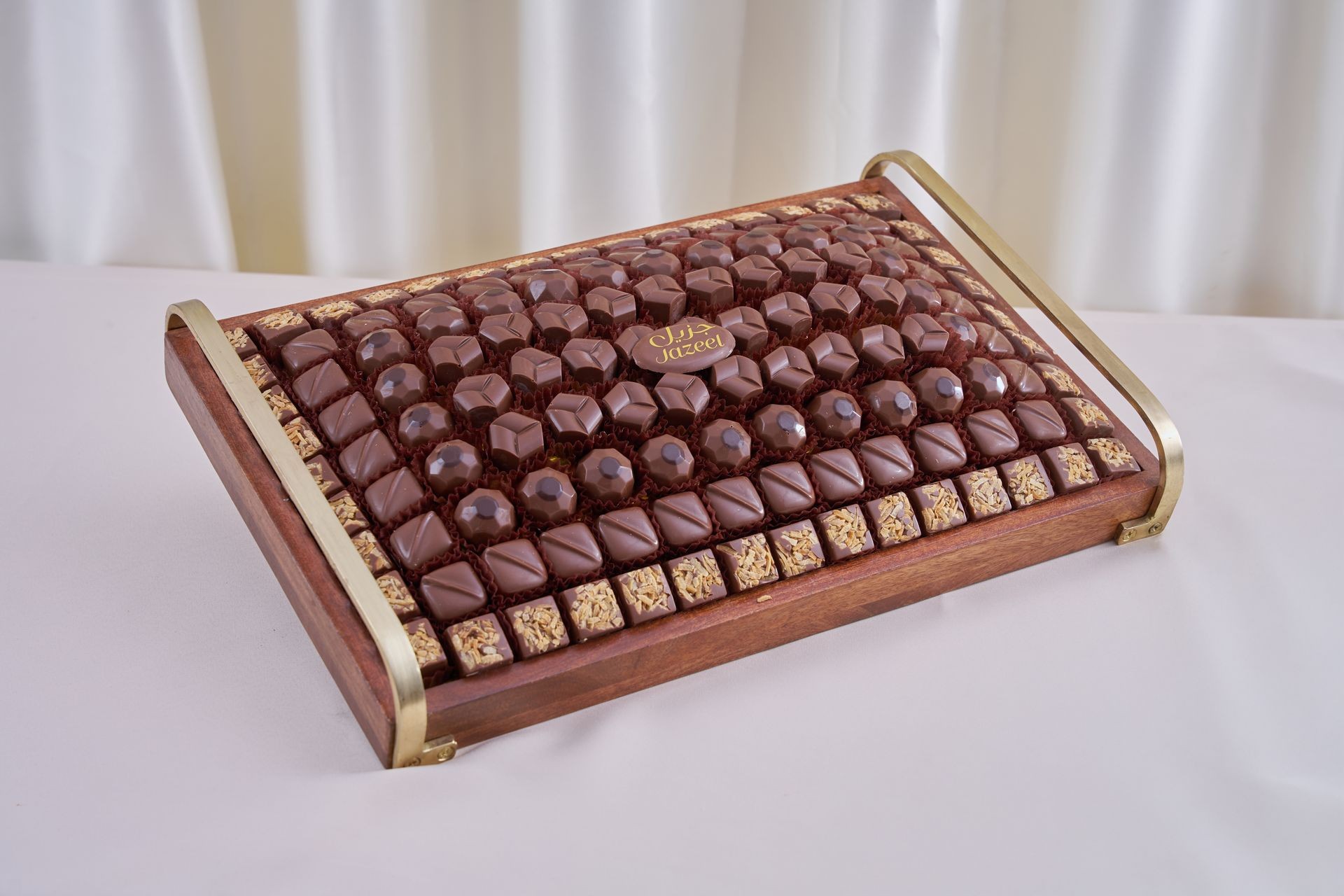 Assorted chocolates arranged on a rectangular wooden tray with gold handles, set against a white background.