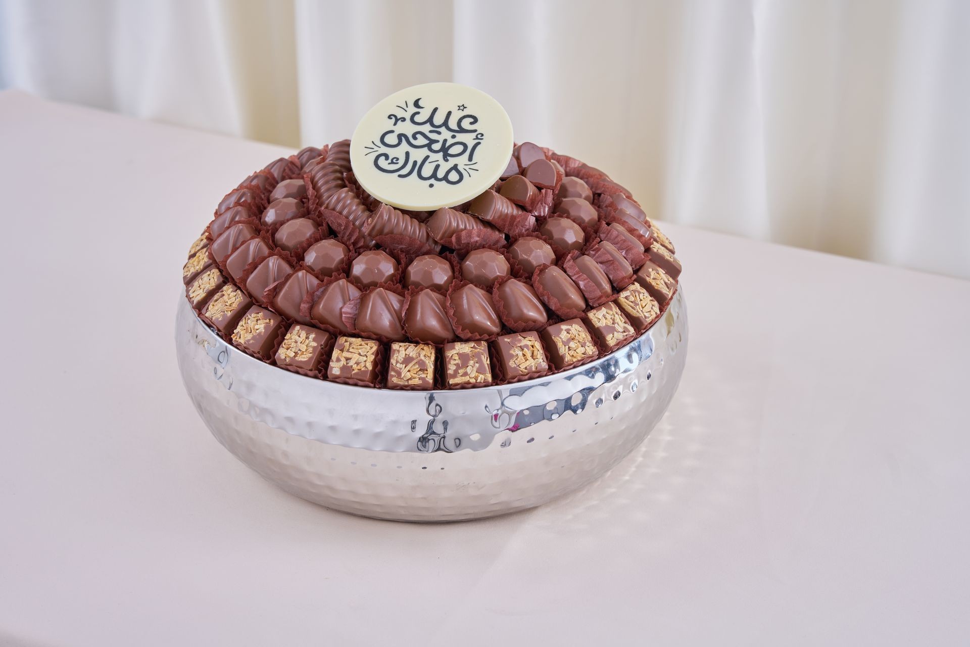 Decorative bowl filled with assorted chocolates, topped with a round white plaque featuring Arabic calligraphy.